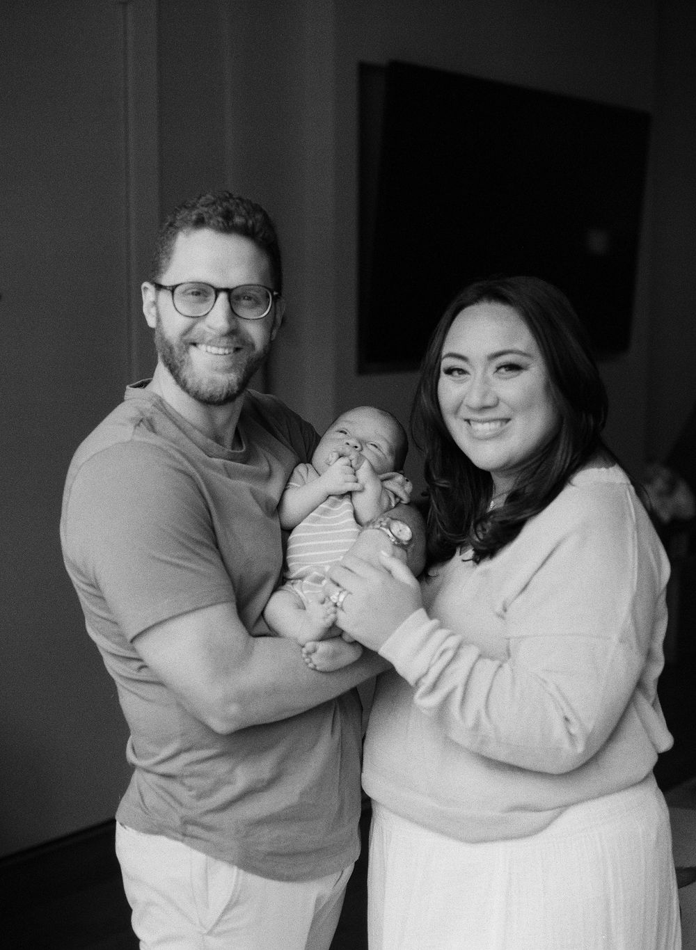 Happy parents smiling while holding newborn baby in black and white family portrait.