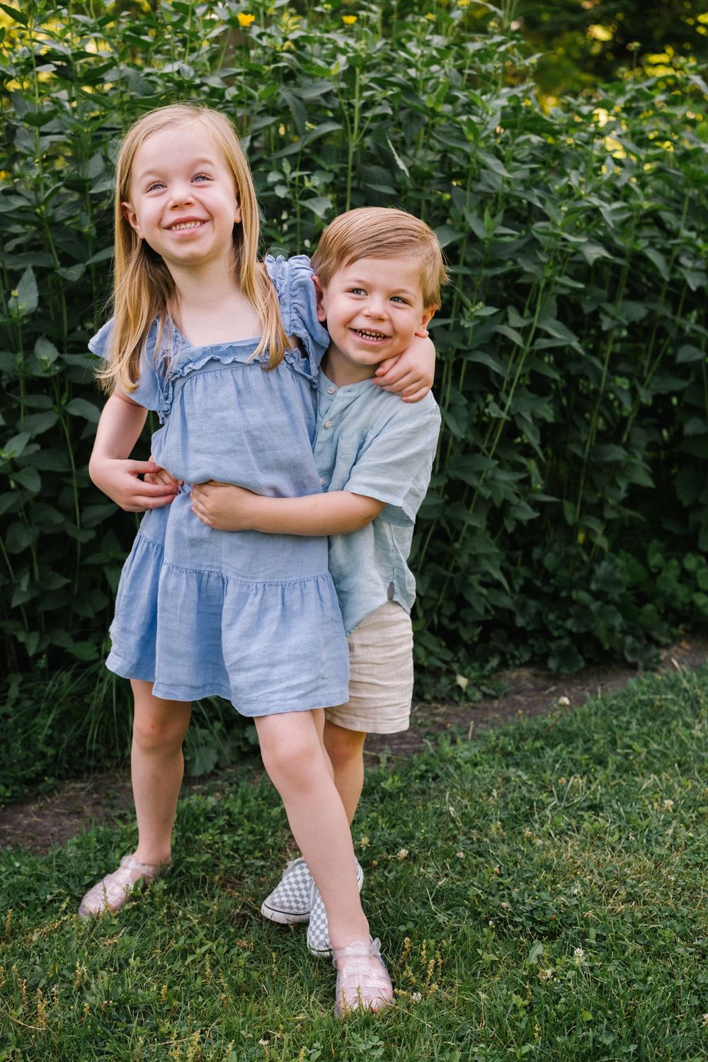 Two young siblings hugging and smiling outdoors in blue outfits, standing on green grass near a leafy hedge.