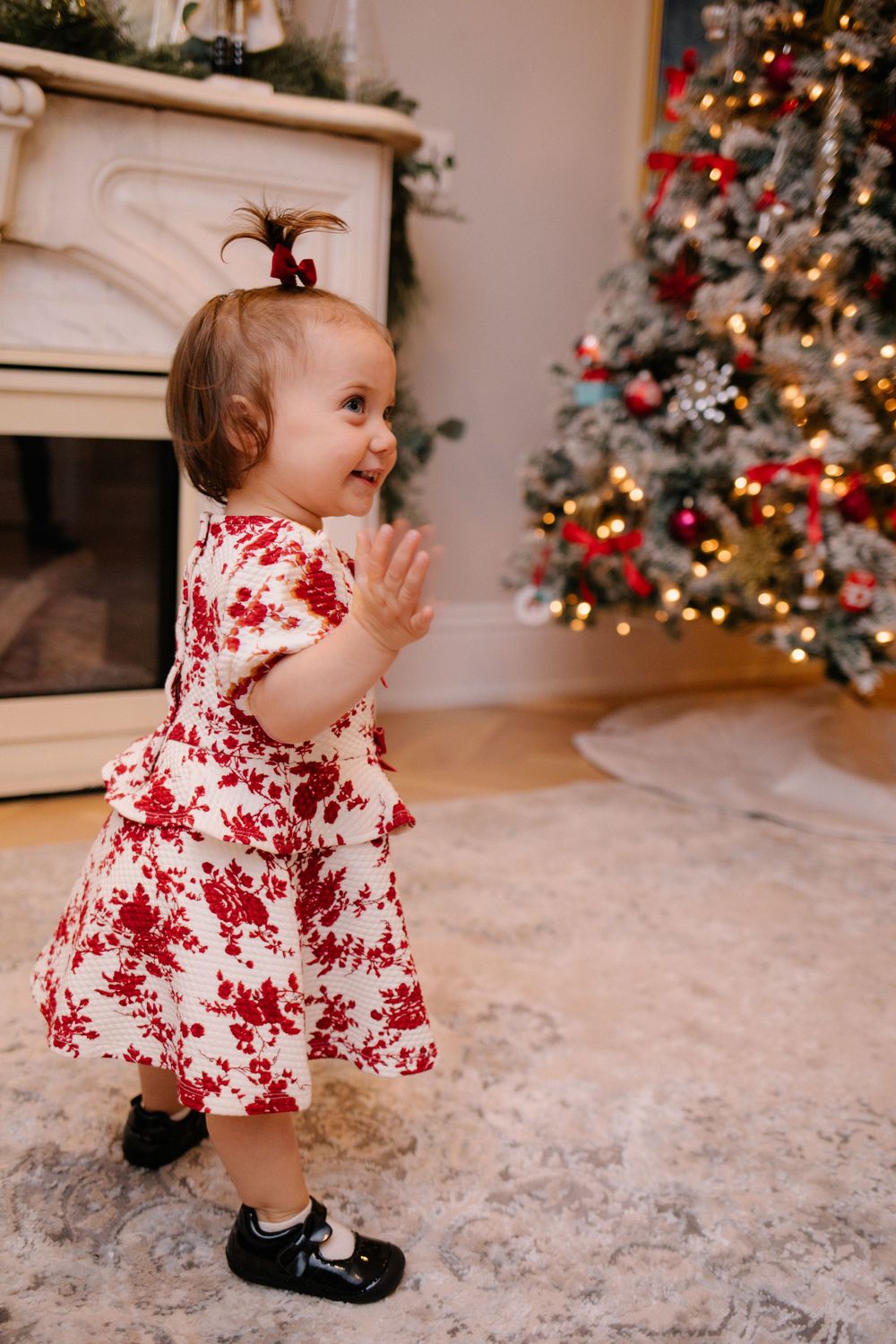 Adorable baby girl in red floral dress clapping near decorated Christmas tree with glowing lights.