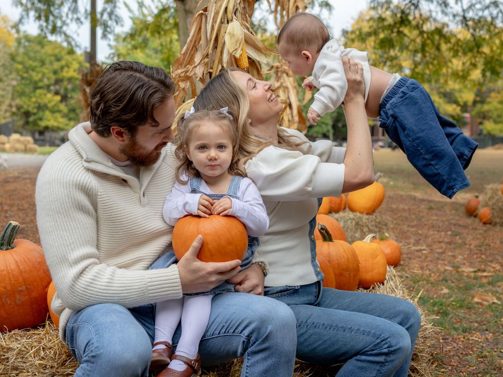 Family enjoying a fall pumpkin patch, parents with toddler girl and baby among orange pumpkins and autumn foliage.