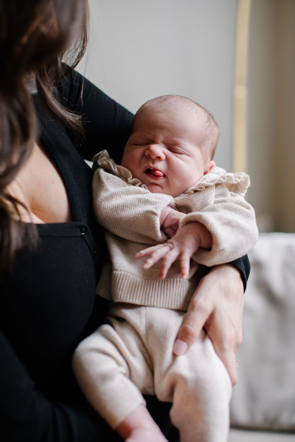 A newborn baby in a beige knit outfit being cradled in the arms of a woman wearing black.