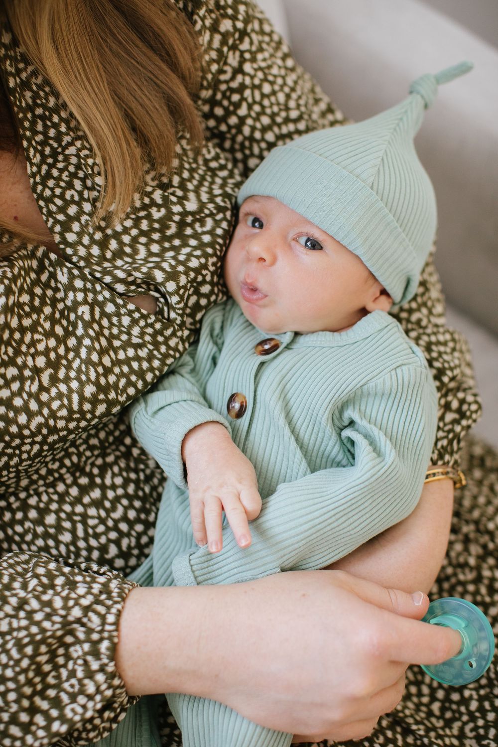 A newborn baby in a sage green ribbed outfit and knotted hat being cradled by a mother in a floral dress.
