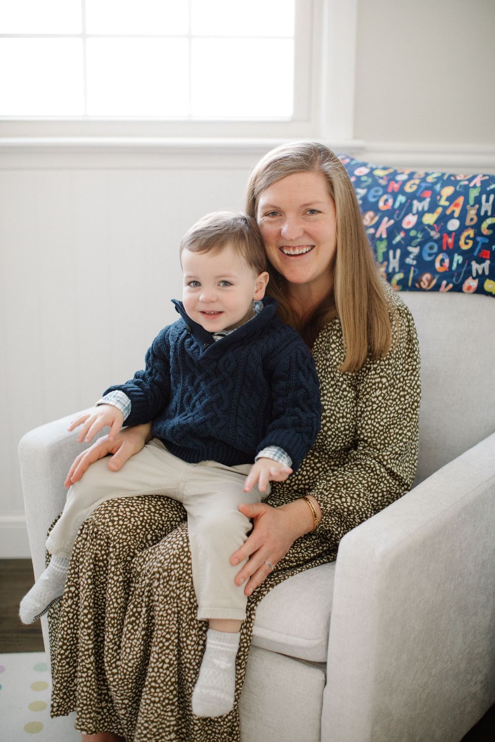 Smiling woman in leopard print dress holds toddler boy in navy sweater on white chair in bright room.
