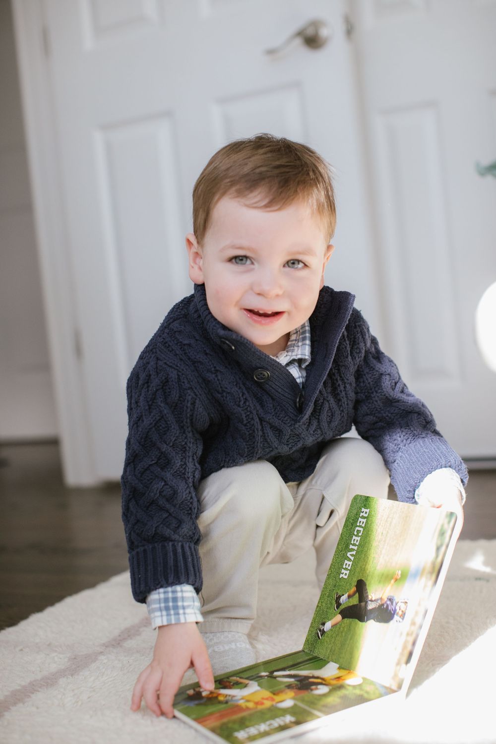 Smiling toddler boy in navy cable-knit sweater sitting on floor holding a colorful picture book.