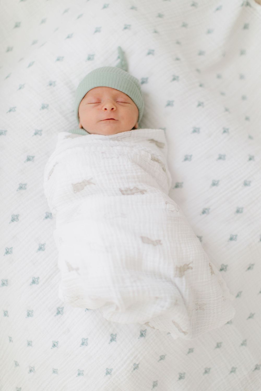 Newborn baby swaddled in white muslin wrap wearing a sage green knit hat, sleeping peacefully on a patterned blanket.