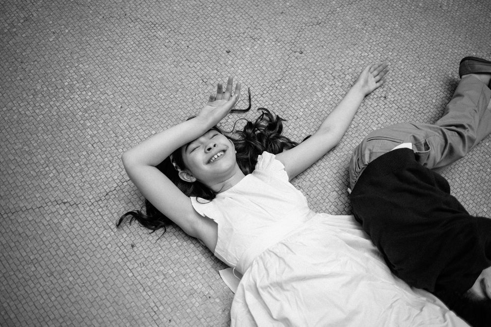 Black and white photo of a smiling girl in white dress lying on ground with arms raised joyfully.