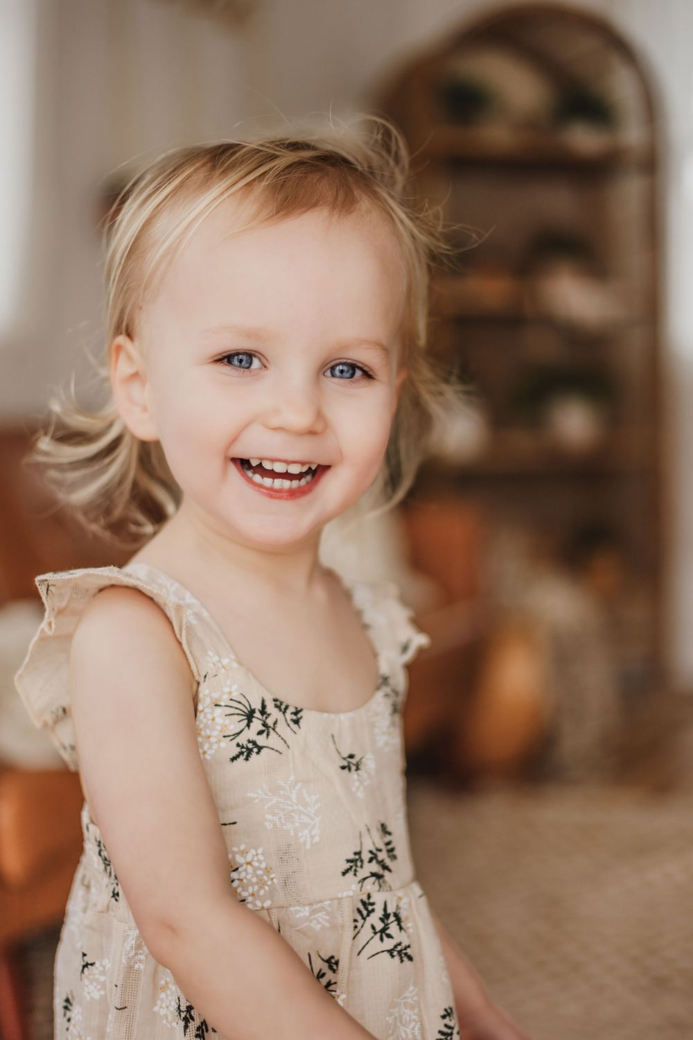 Toddler in floral dress twirls and smiles against warm indoor background.