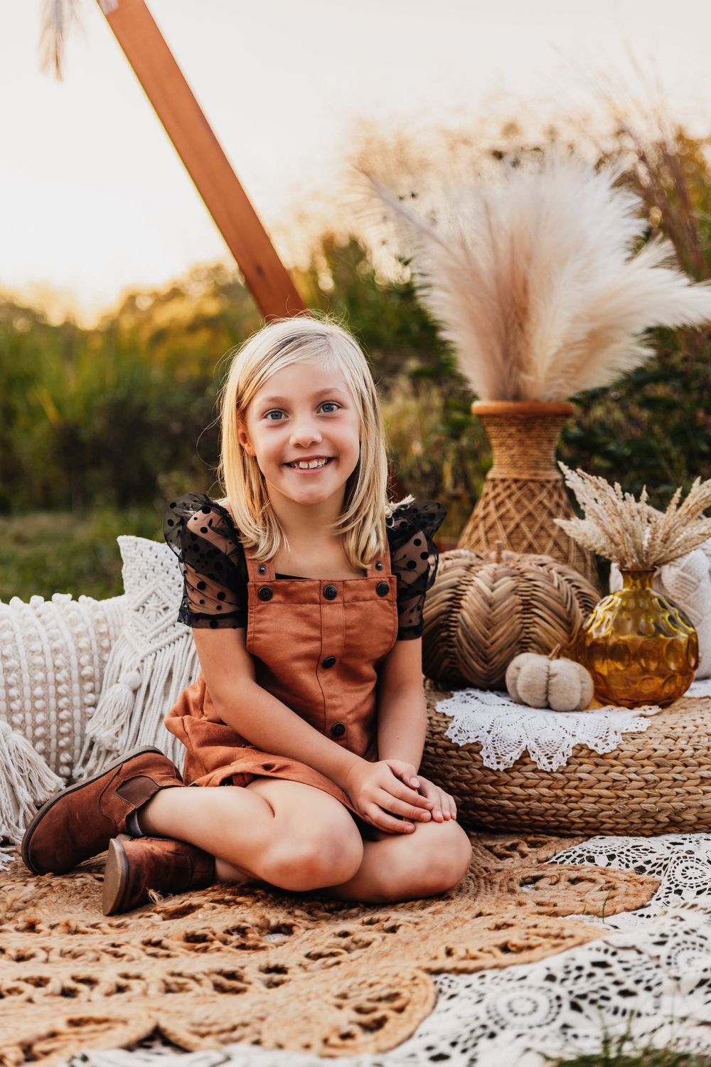 A child sits on a patterned blanket with boho decor and pampas grass arrangements.