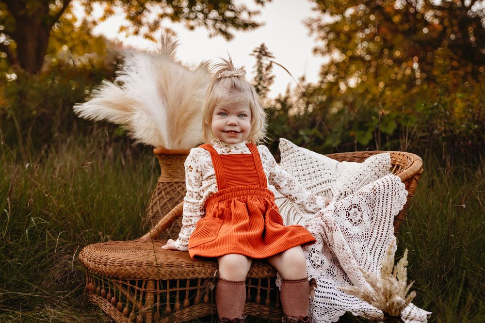 A child in a red dress sits on a wicker basket with decorative pampas grass and lace in an outdoor setting.
