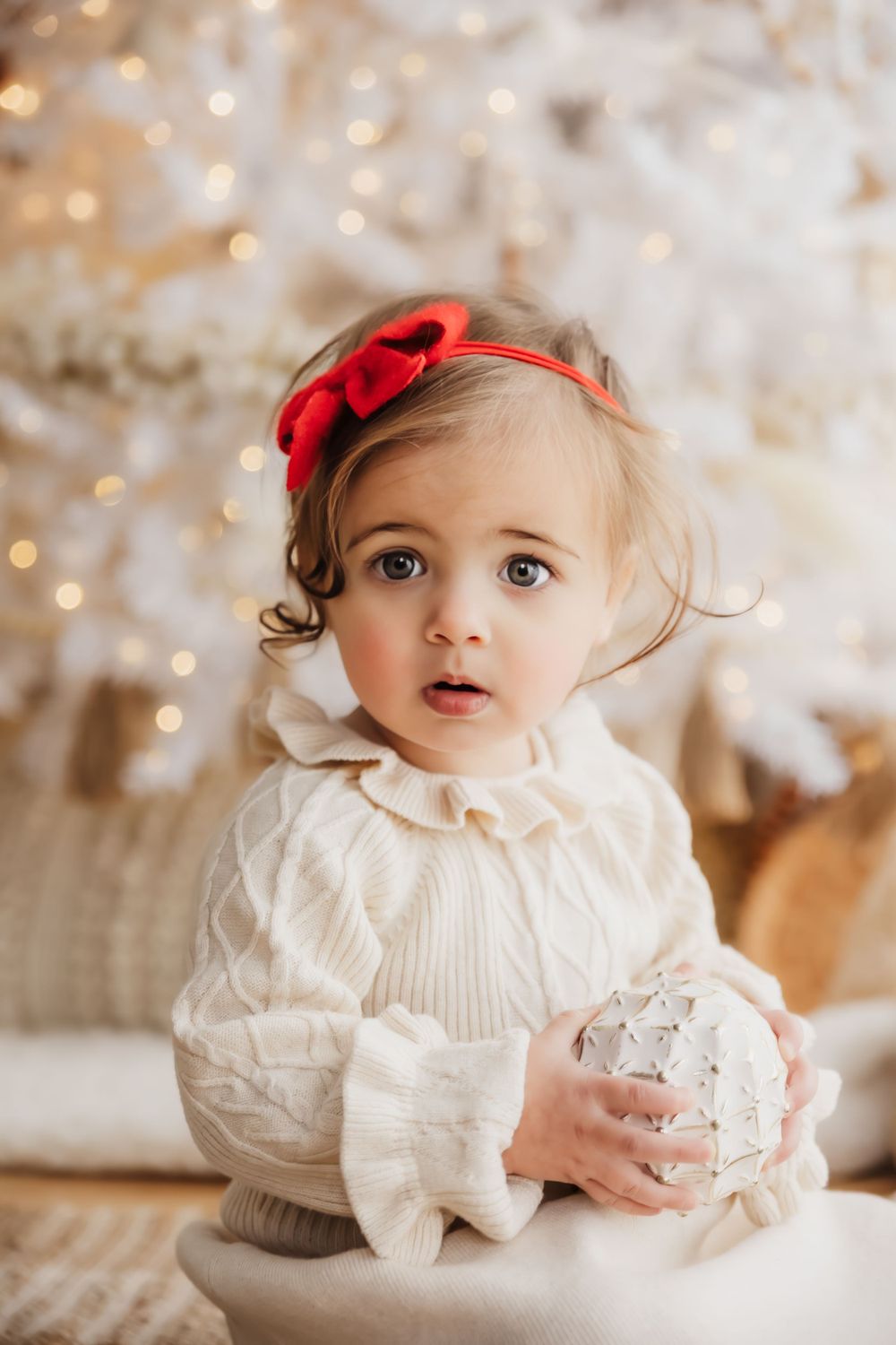 Baby in white sweater holding ornament with red bow headband against holiday backdrop.