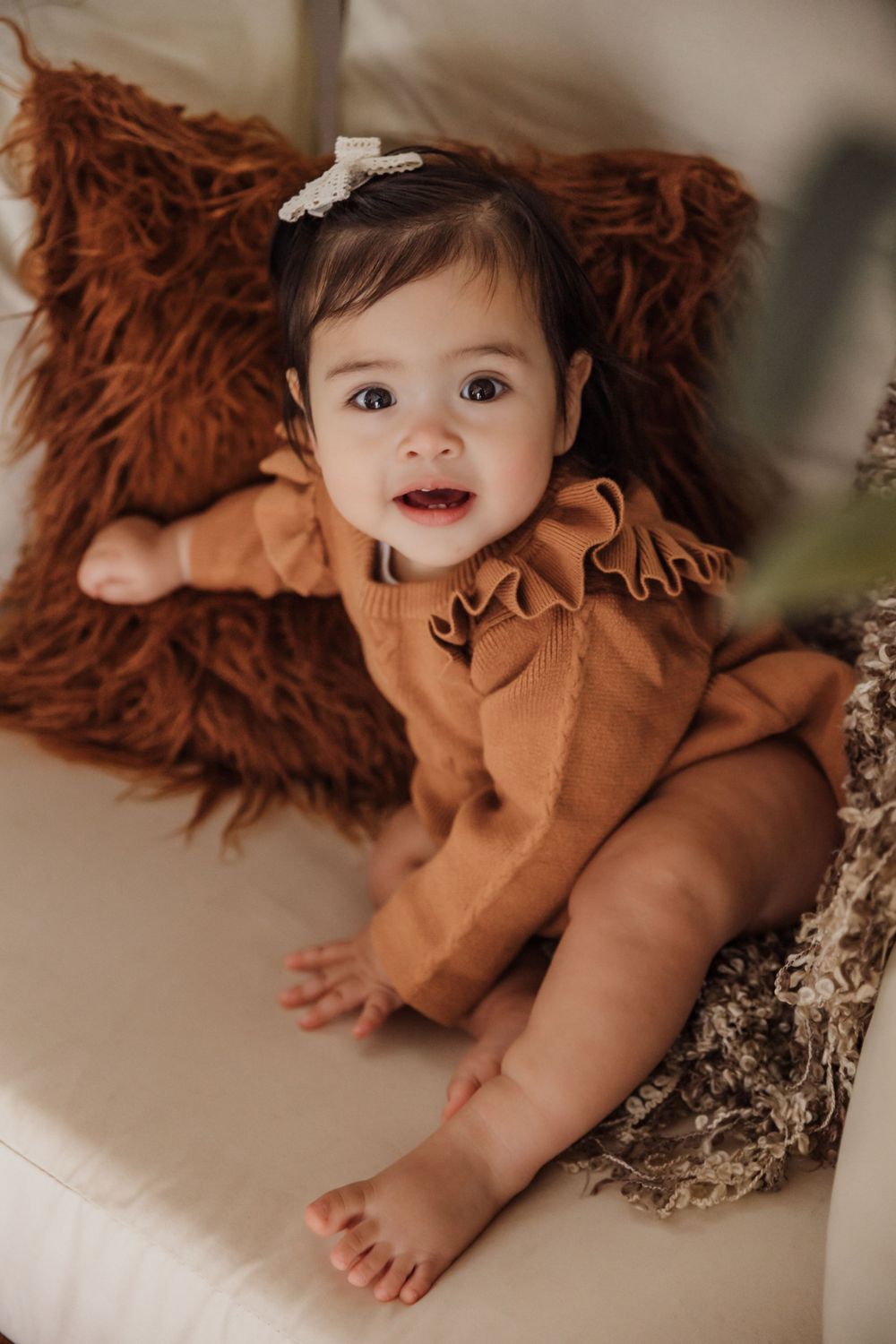 Baby in rust-colored outfit smiling while lying on fuzzy brown pillow.