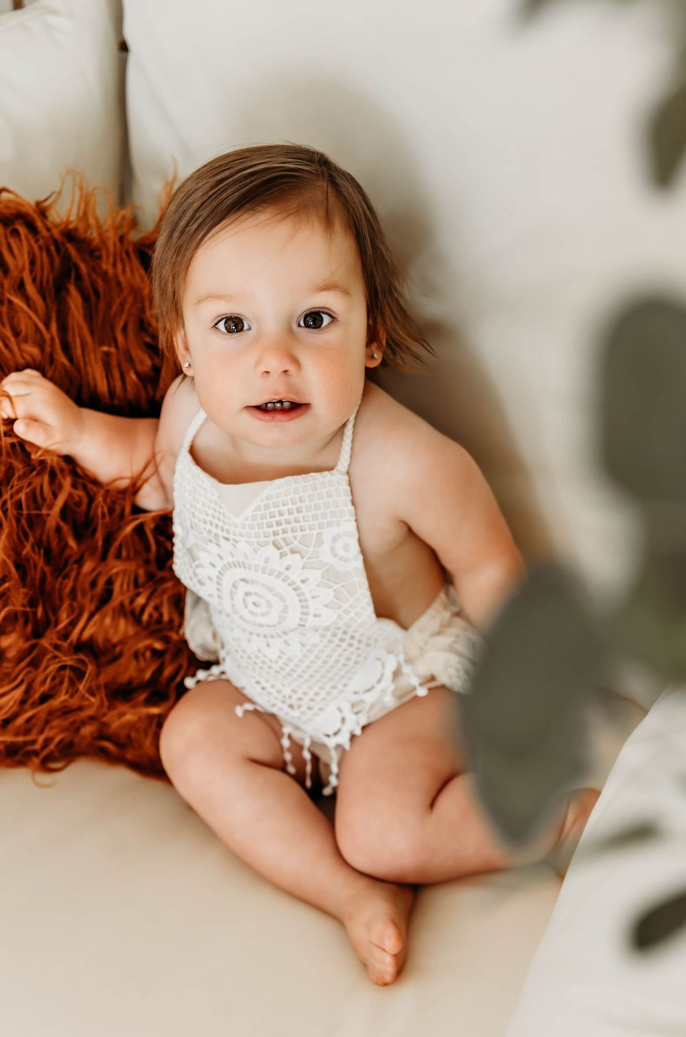 Baby wearing white lace romper poses on orange textured rug.