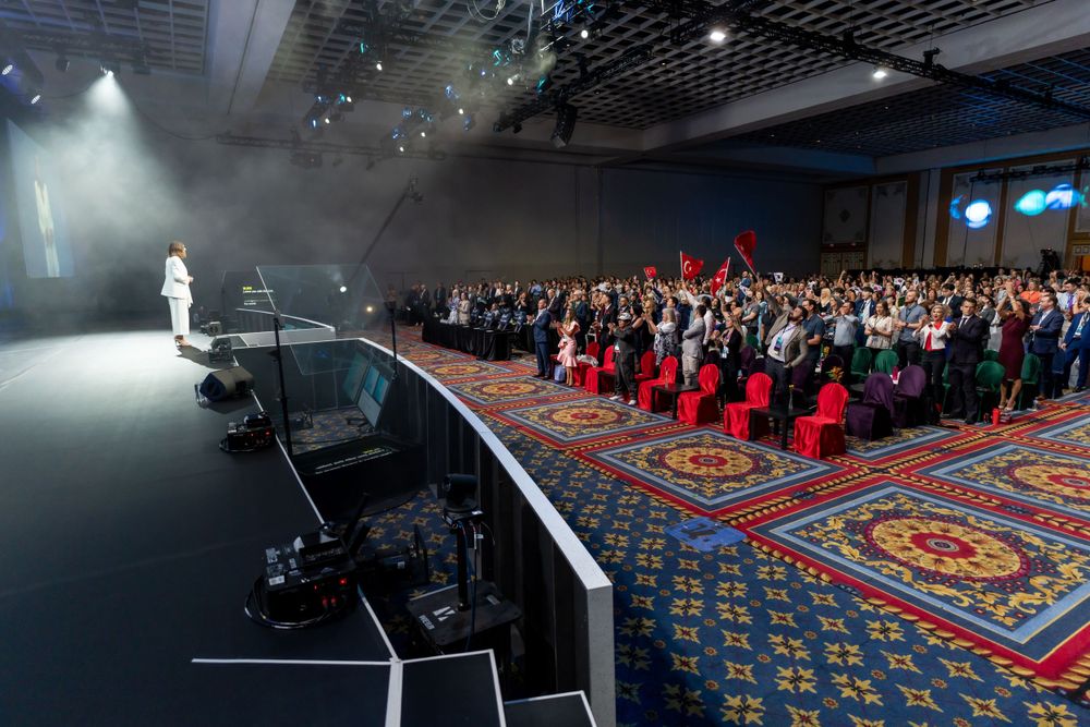 View of a crowded convention hall with a lit stage and audience seated on patterned carpet.