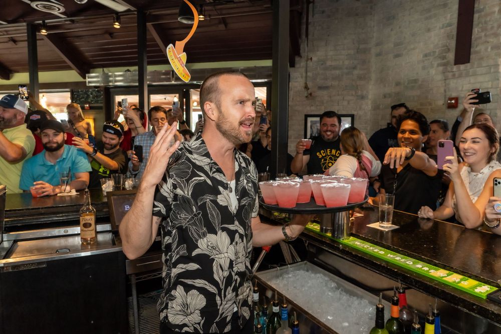 Bartender in floral shirt serving drinks at a crowded bar counter.