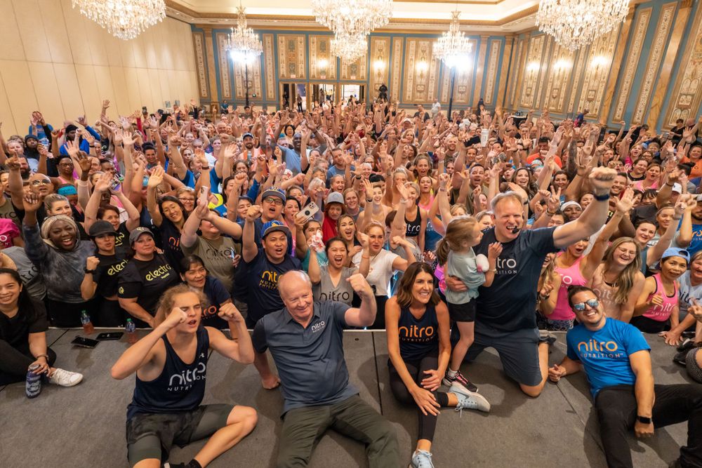 Enthusiastic crowd in formal attire posing with raised arms in an ornate ballroom with crystal chandeliers.
