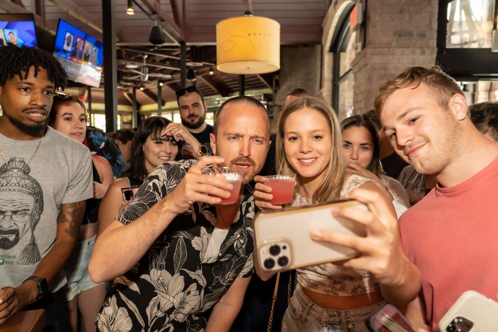 Group of patrons enjoying drinks and taking selfies at a lively bar.