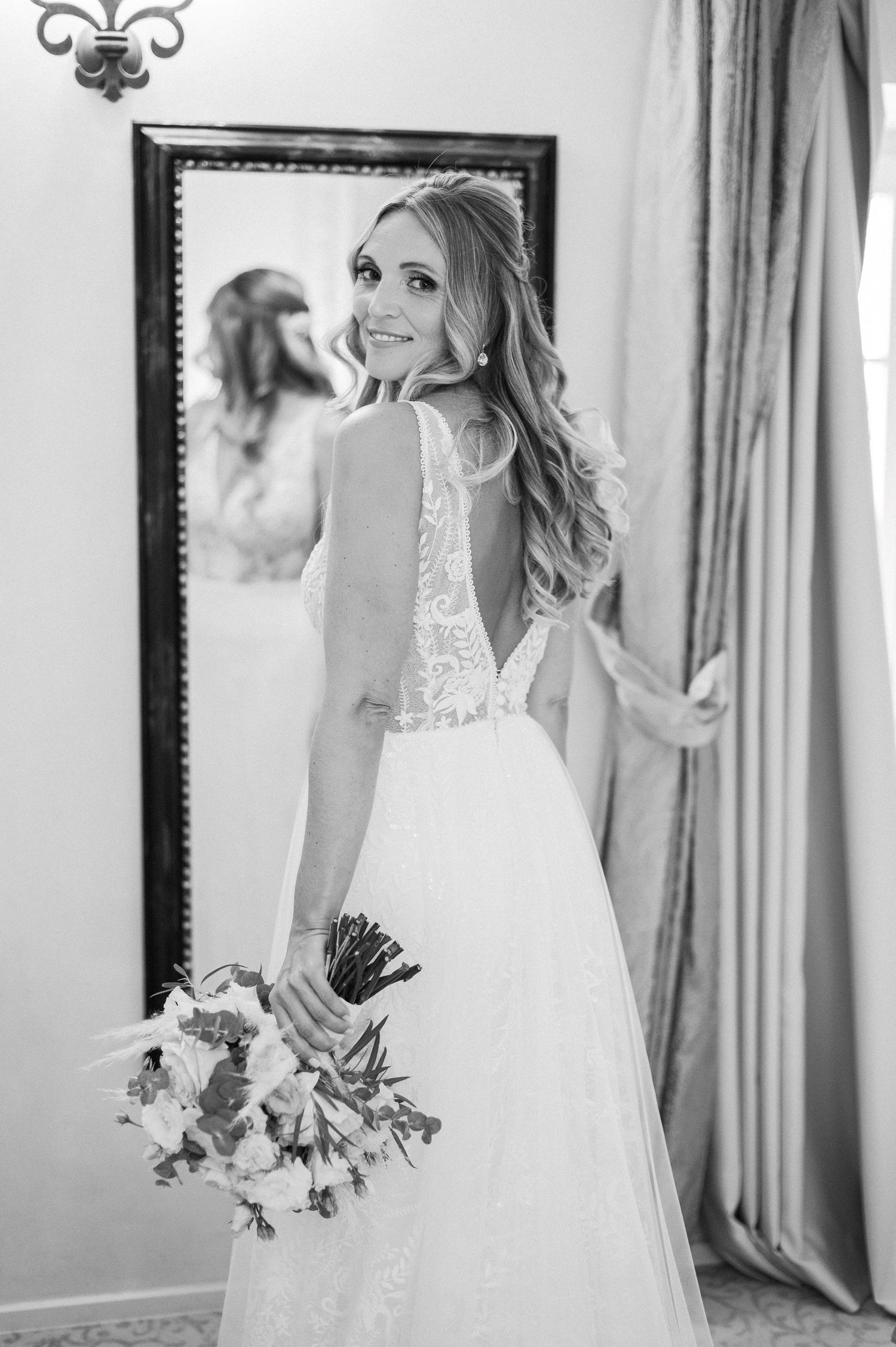 Bride in elegant lace back wedding dress poses with bouquet in front of mirror.