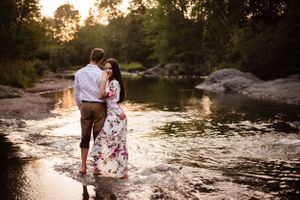 A couple stands together in shallow water during golden hour near a rocky shore.