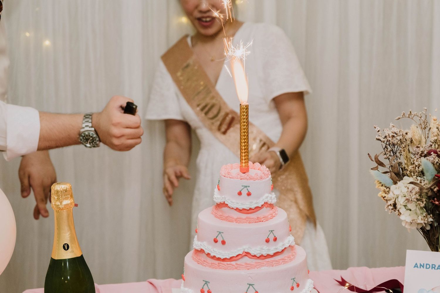Someone lighting a candle on a pink tiered birthday cake with champagne nearby.