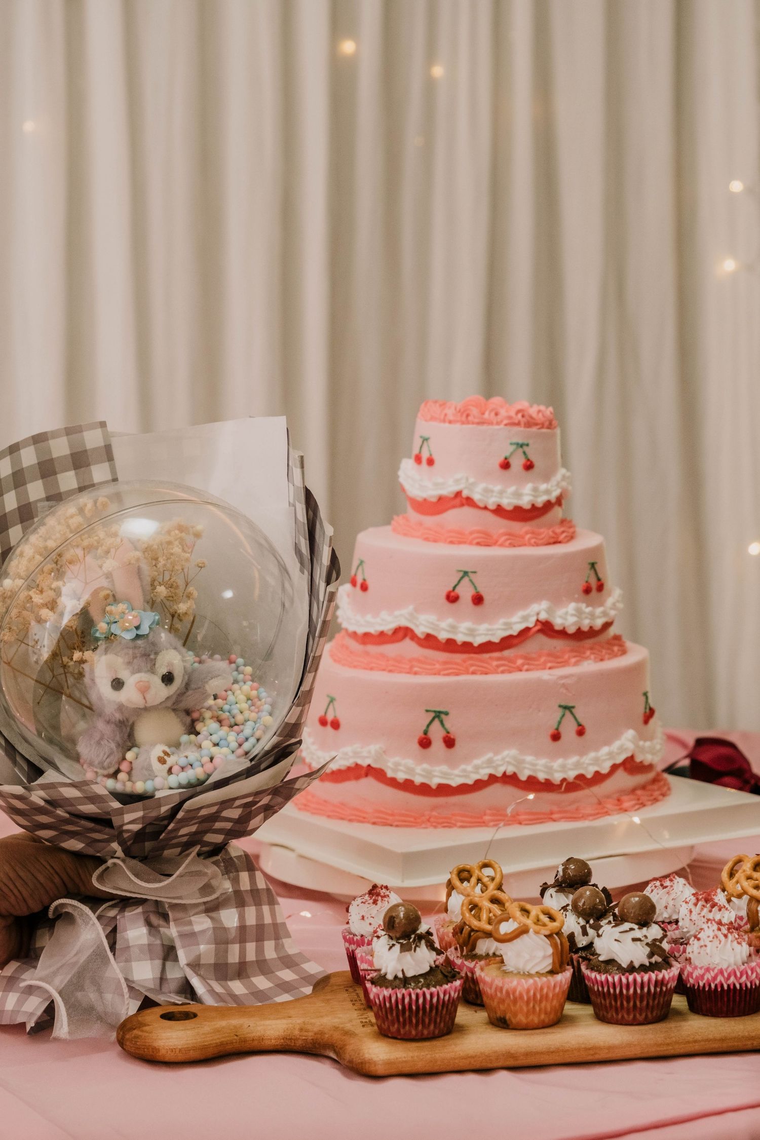 Three-tiered pink and white cake with cherry decorations alongside cupcakes and party decor.