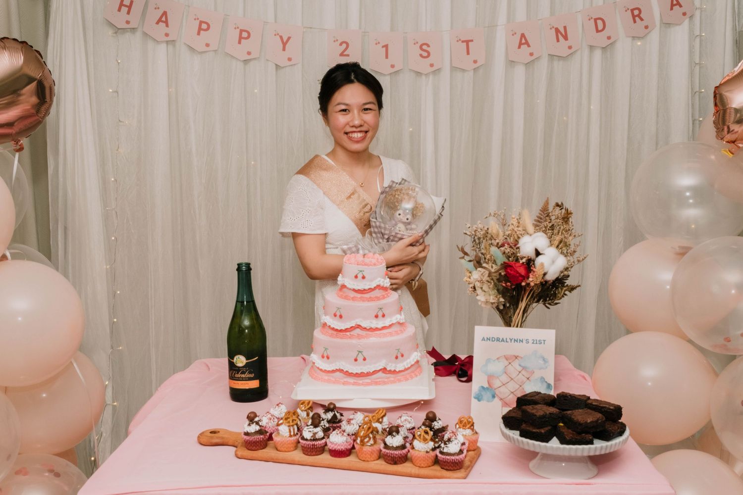 A festive birthday setup with pink and white balloons, a tiered cake, and champagne.
