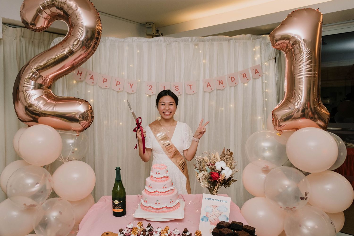 A birthday celebration setup with rose gold number balloons, pink decor, and a tiered cake.