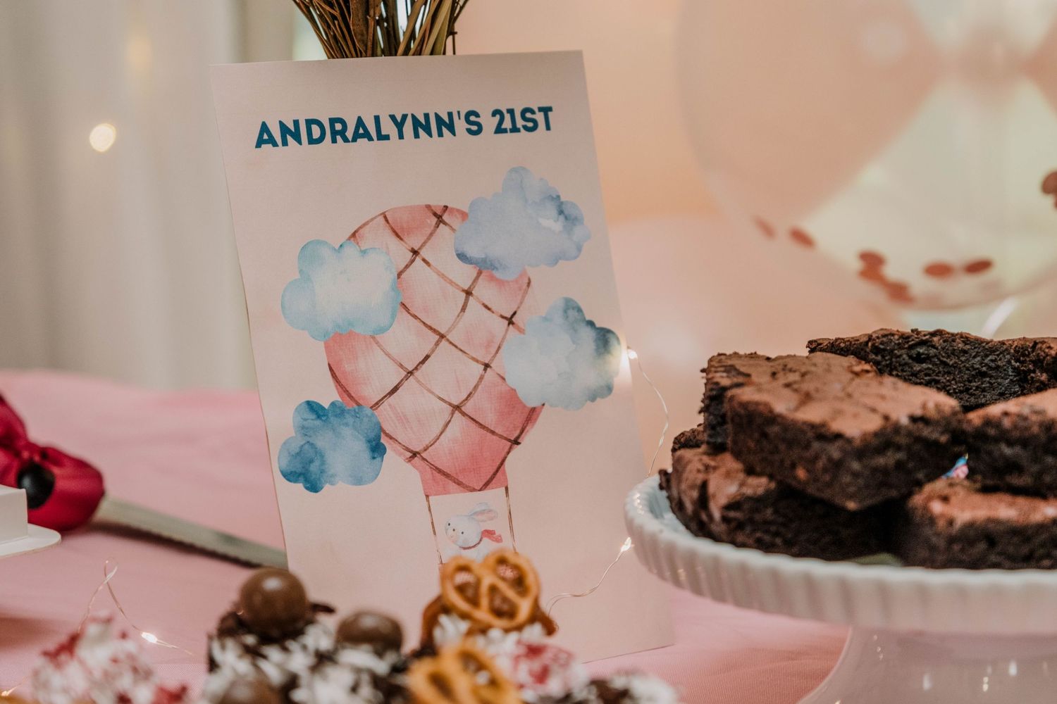 Decorative party setup with Andralynn's 21st signage, clouds design, and brownies on a pink tablecloth.