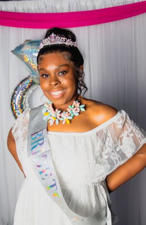 Woman in white dress wearing tiara and sash smiling at camera.
