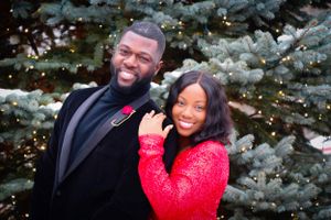 A couple smiles together in front of a snowy evergreen tree with twinkling lights.