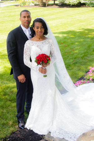 Bride in lace wedding gown holds red bouquet while posing with groom on manicured lawn.