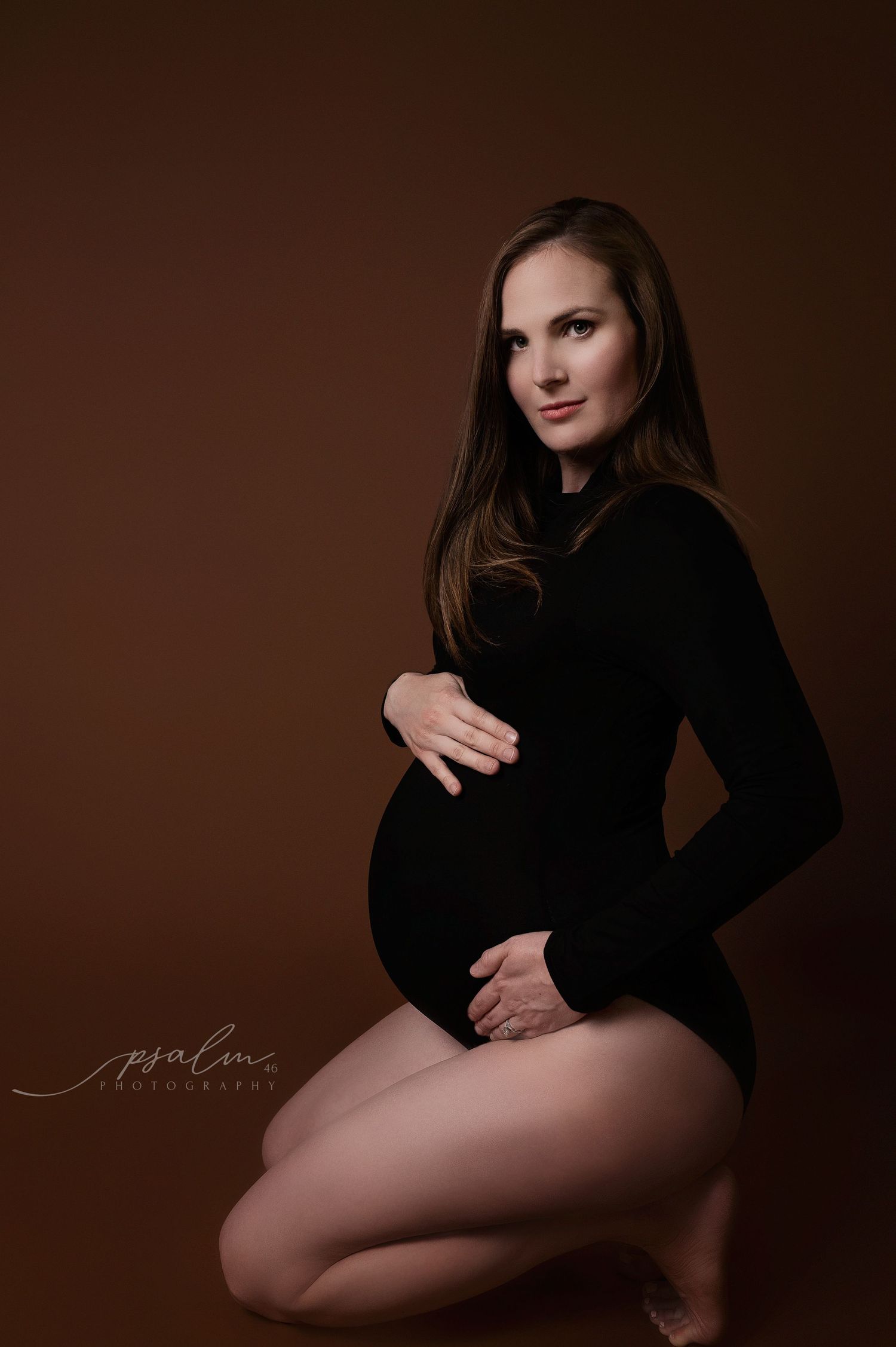 Artistic maternity session with woman in black against dark brown background.