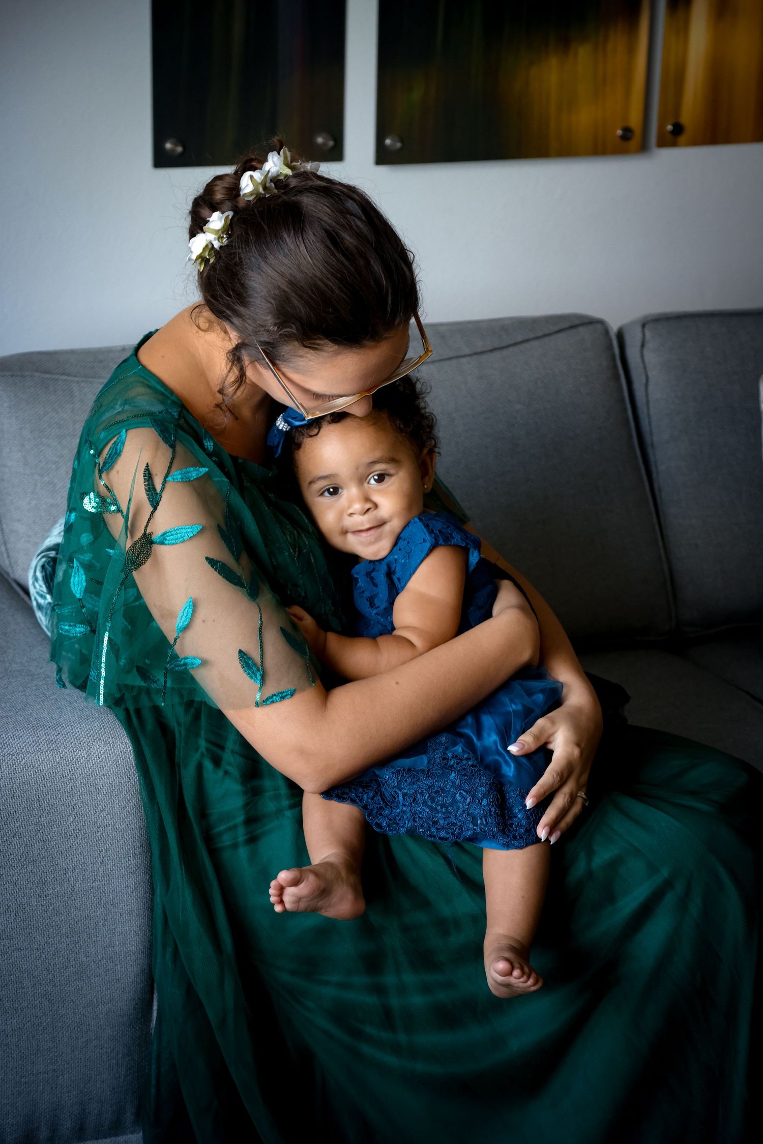 Person in emerald green dress embraces small child wearing blue dress on grey couch.