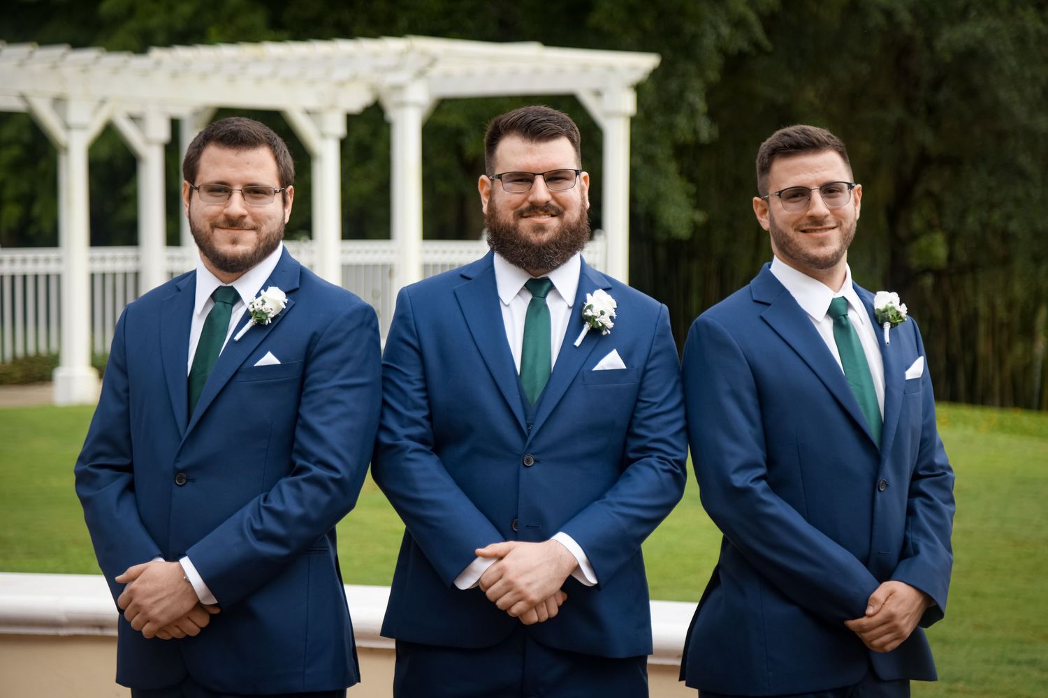 Three groomsmen in matching navy suits stand together in front of a white garden gazebo.