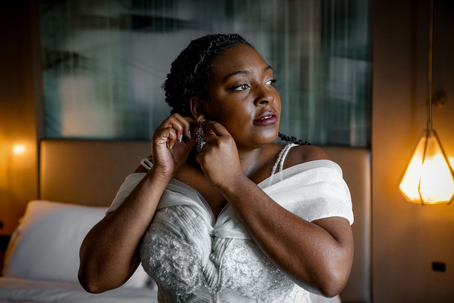 Person in white dress puts on earrings while getting ready in warm lighting.