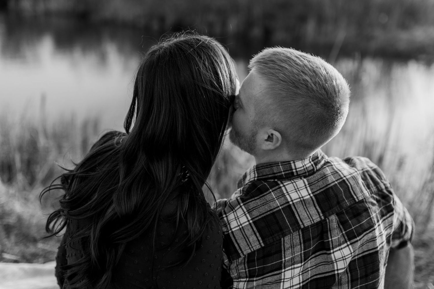 A man kisses his fiance's cheek as they sit looking at a pond in a tall grass field during an engagement photo session.