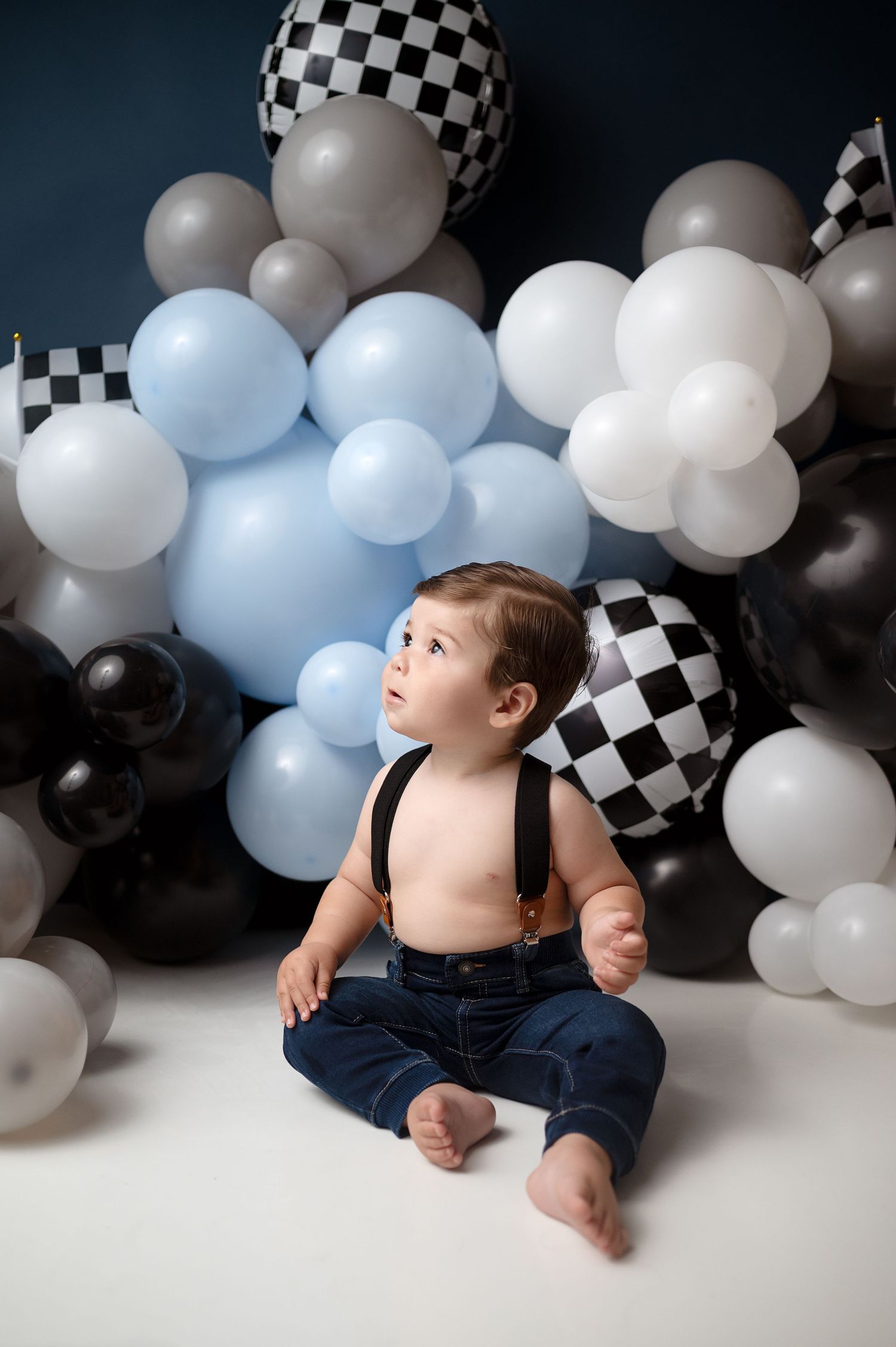 Toddler in jeans looking up while sitting against blue and checkered balloon display.