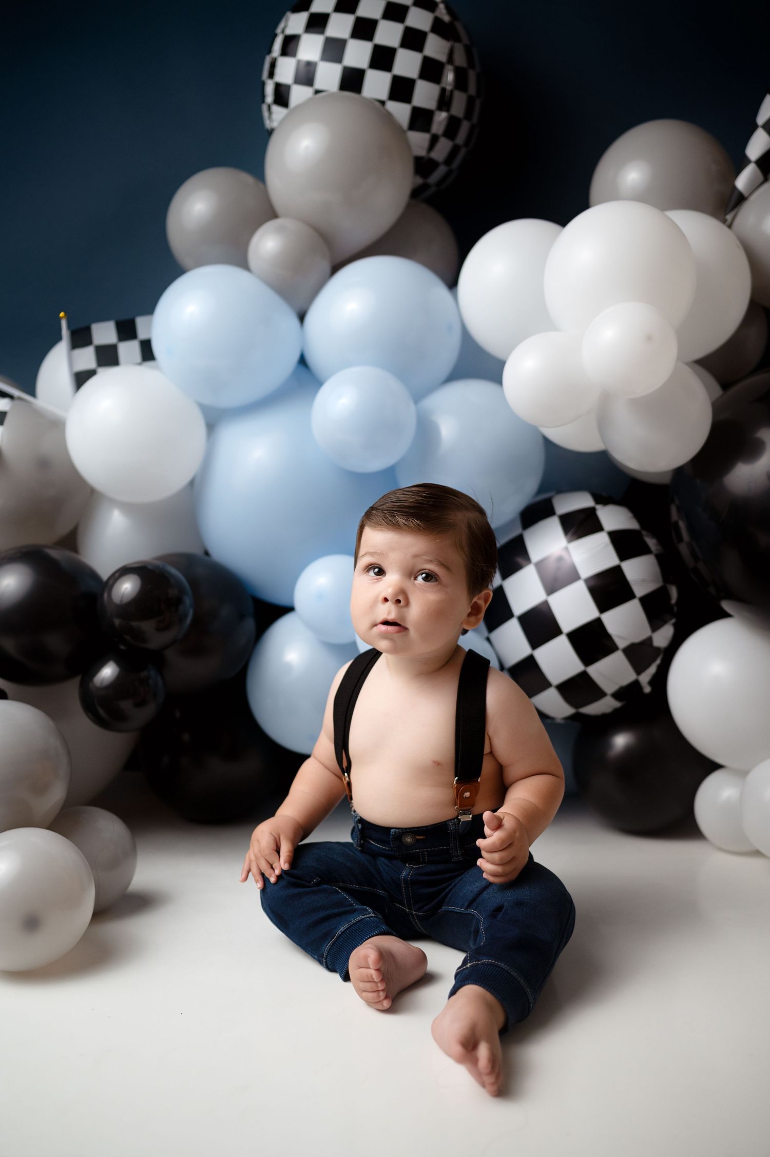 Child in suspenders sitting against decorative balloon wall with checkered patterns.