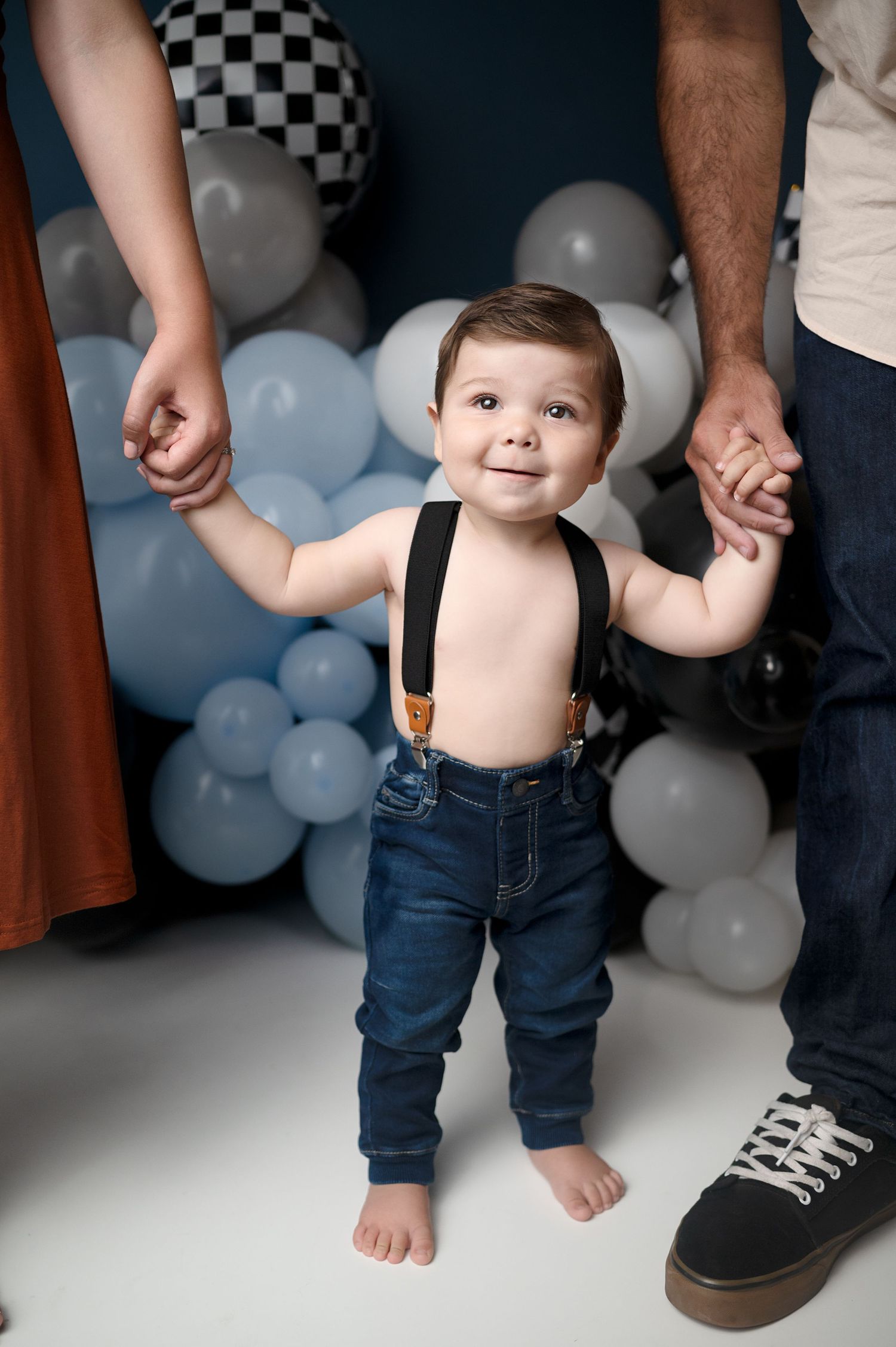 Toddler in suspenders and jeans holding parents hands against balloon backdrop.