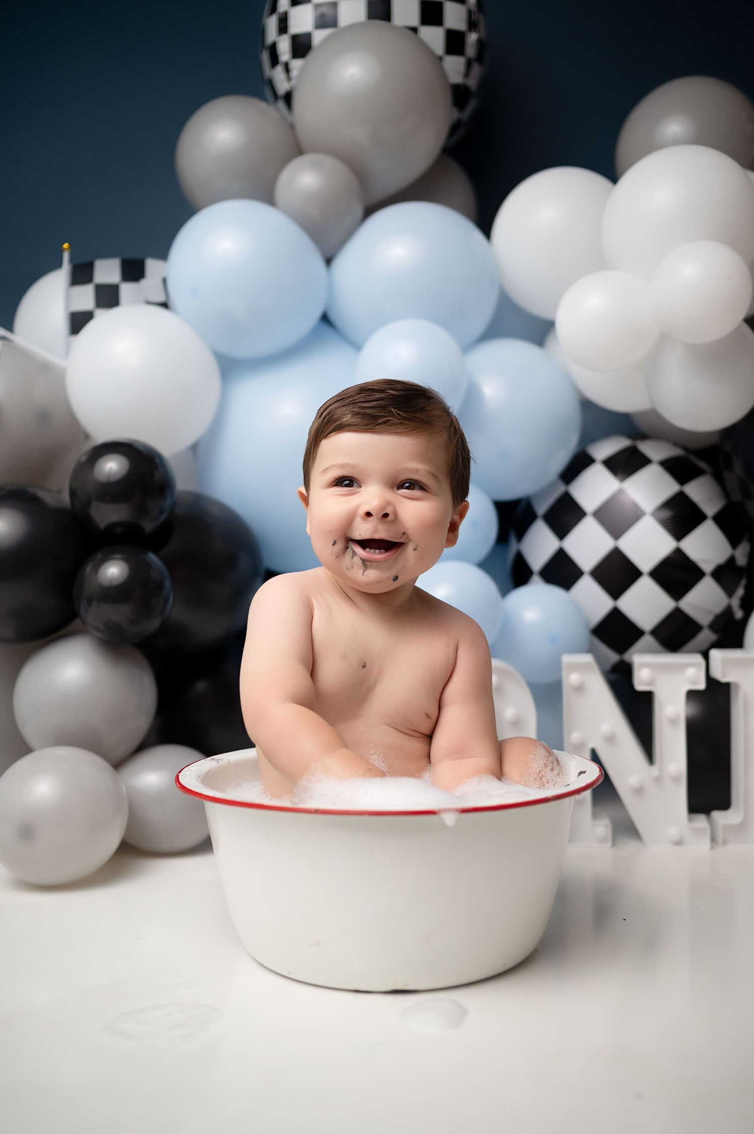 Baby sits in white bowl against racing-themed balloon backdrop for milestone photo.