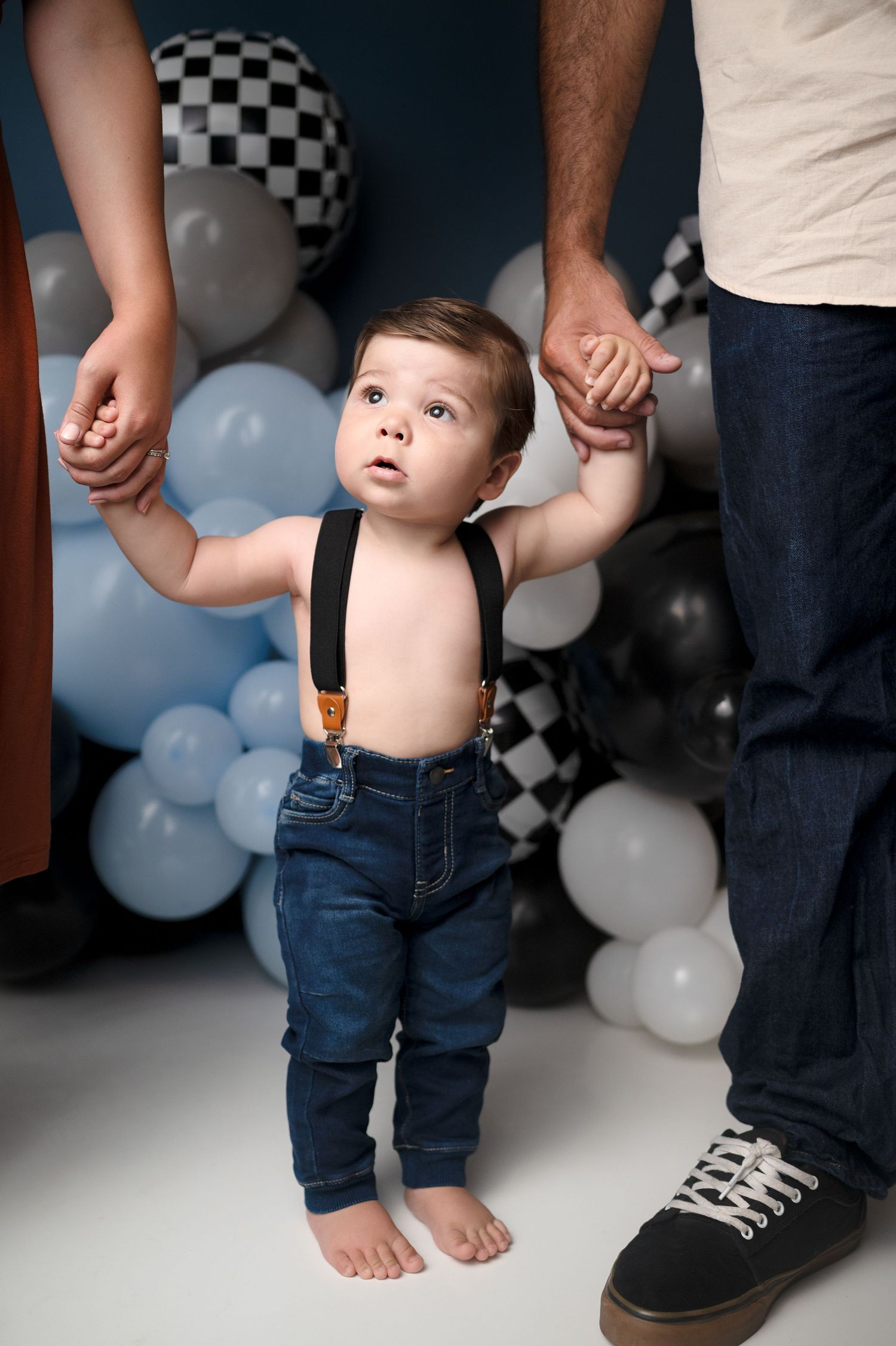 Side view of toddler in suspenders holding hands with parents against decorated balloon wall.