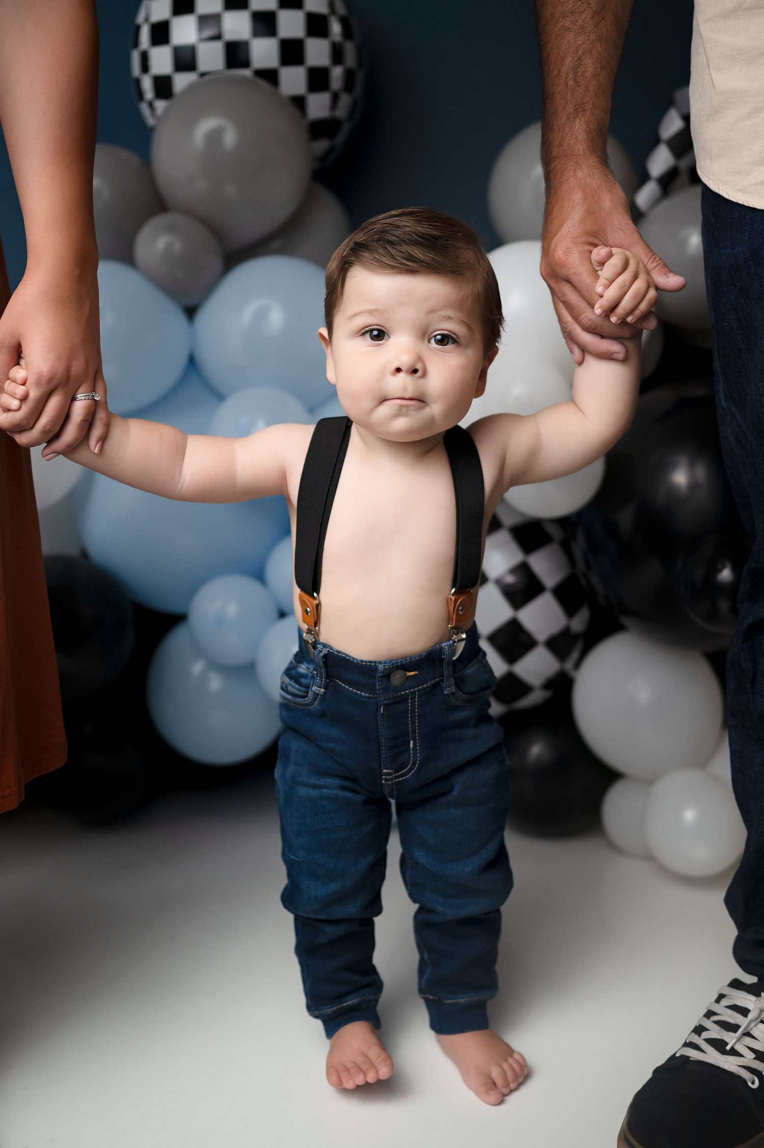 Toddler in suspenders and jeans standing with balloon party backdrop.