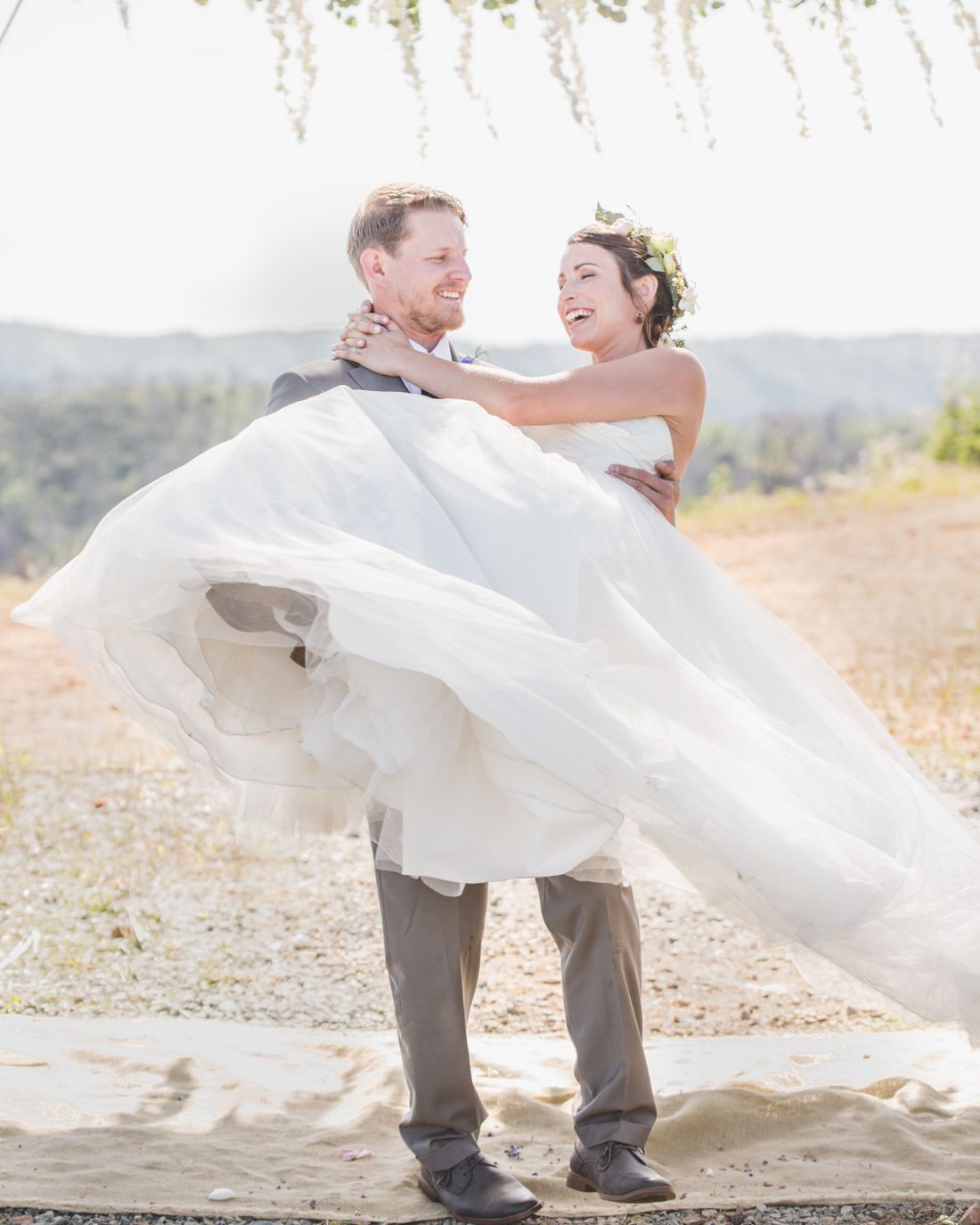 A groom lifts his bride in a white flowing dress at their outdoor wedding.