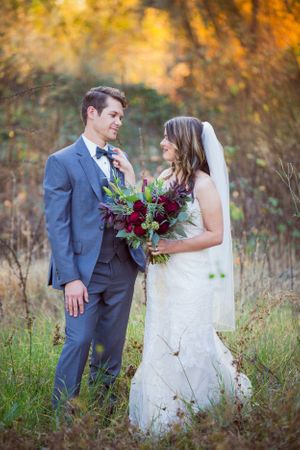Bride in white dress holds burgundy bouquet while standing with groom in autumn field.