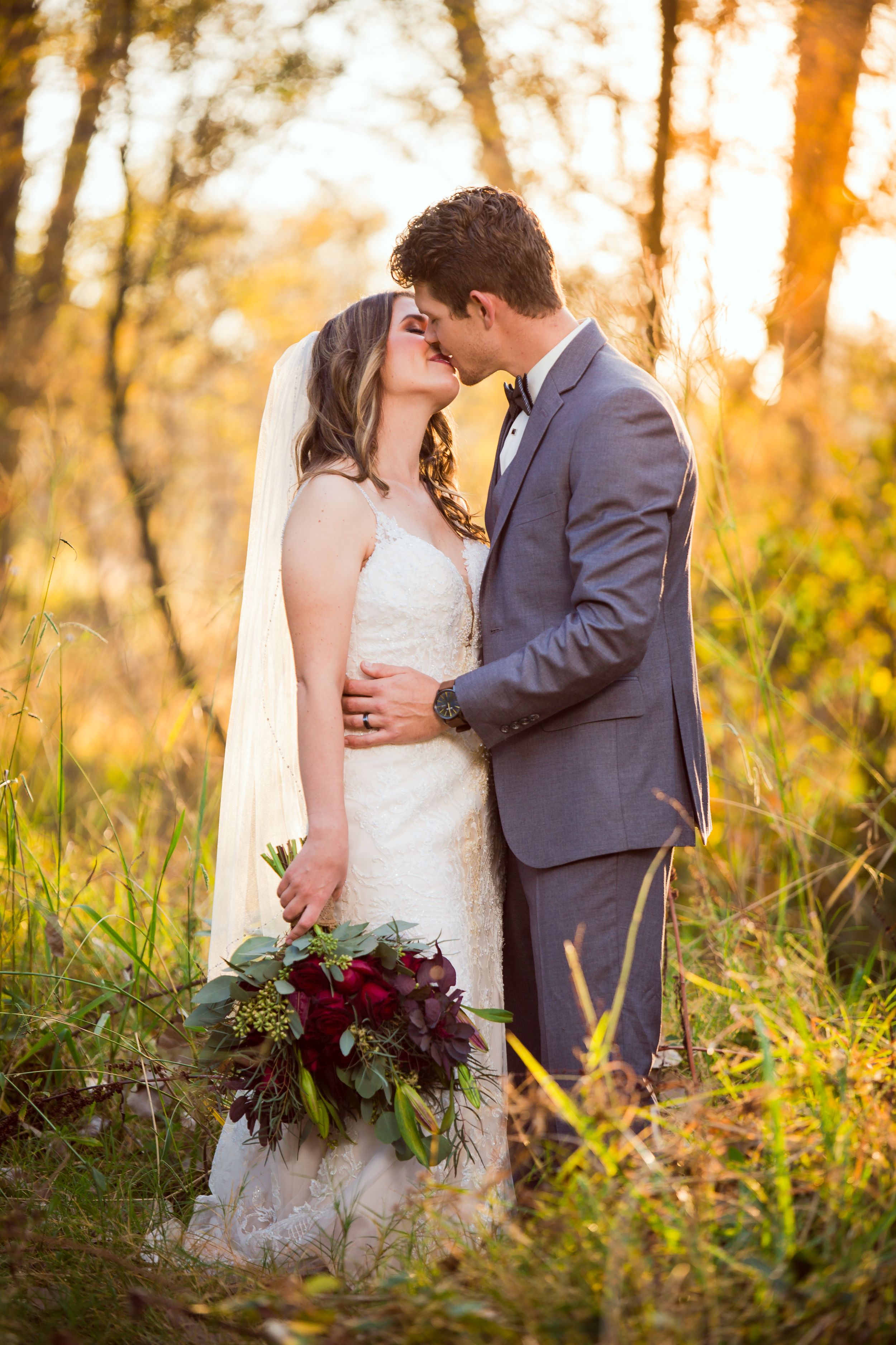 Newlyweds kiss while holding burgundy bouquet in sunlit field.