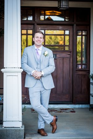 Groom in light grey suit poses confidently on porch in front of wooden door.