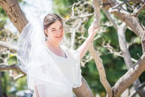 Bride in flowing white veil poses joyfully among tree branches.