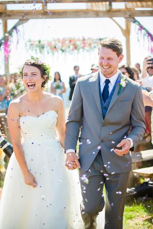 Joyful couple exits their wedding ceremony under confetti shower at rustic outdoor venue.