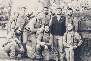 Group of groomsmen in vintage-style attire pose together by garden fence.