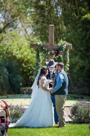 Wedding ceremony under floral-decorated cross arch in garden setting.