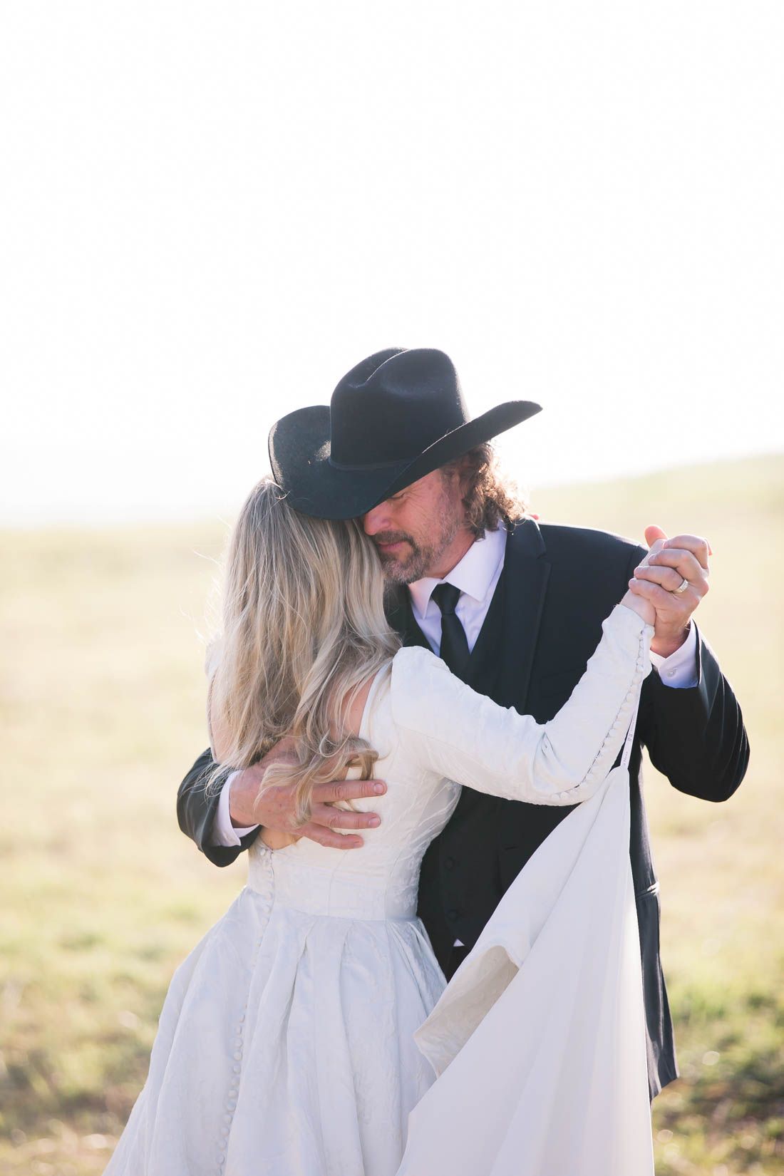 A couple in wedding attire dancing together in a field, with the groom wearing a black cowboy hat.